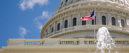 Close-up view of the United States Capitol building with an American flag flying in front of it. The dome of the Capitol is prominently featured, and there is a fountain in the foreground. The sky is clear with a few scattered clouds.