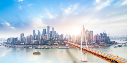 A panoramic view of a modern city skyline with numerous high-rise buildings under a bright, partly cloudy sky. In the foreground, an orange and white suspension bridge spans across a wide river, leading towards the city situated on both sides of the river. Several boats are visible on the water.