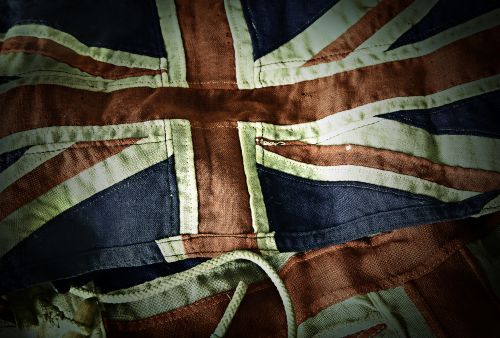 A close-up of a worn and slightly tattered Union Jack flag, which is the national flag of the United Kingdom. The fabric appears aged and frayed at some edges, with visible stitching and folds. The colors are somewhat faded, indicating that the flag has been used or exposed to elements for an extended period.