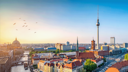 Panoramic view of Berlin cityscape during daytime, featuring historic and modern buildings, with the Fernsehturm Berlin (Berlin TV Tower) prominently in the center, clear sky with birds flying on the left, and the Spree River winding through the