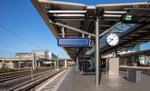 A train station platform with tracks on the left side. There is a large clock mounted on a pole, displaying the time as approximately 10:10. Above the clock, there is an empty digital display board. The platform has a roof structure with beams and lights, and there are signs indicating directions, including an emergency exit sign with a green arrow pointing to the right. In the background, there are additional platforms and railway infrastructure visible under a clear blue sky.