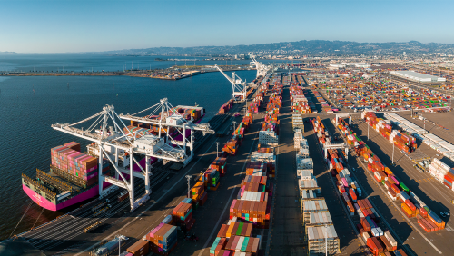 Large port with numerous shipping containers and cranes. Several container ships are docked at the port, with cranes actively loading or unloading containers. The port is situated by a body of water, and there are many rows of colorful shipping