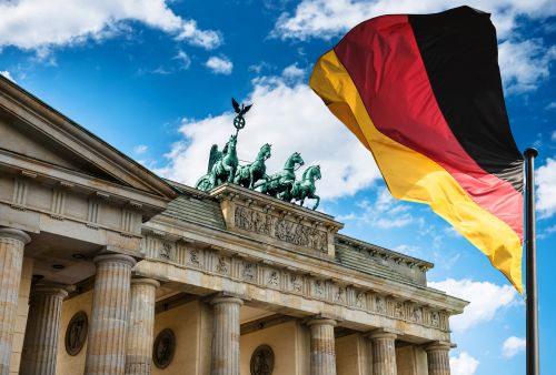 Brandenburg Gate in Berlin, Germany, with a large German flag waving prominently in the foreground. The sky is blue with scattered clouds.